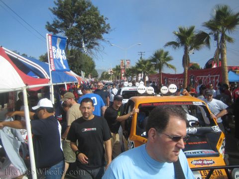 1990 Ford Bronco - dense crowd packs a palm-lined street at an off-road racing event, with PIAA-marked race vehicles and Baja 1000 banners.