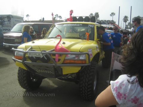 1990 Ford Bronco - yellow off-road race truck marked '740' with a pink flamingo and a 'Beware of Attack Flamingo' sign sits at an event with onlookers.