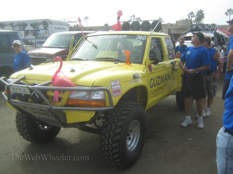 1990 Ford Bronco - yellow off-road race truck '740' lettered 'Guzman Construction' with pink flamingos sits among a crowd in blue shirts.