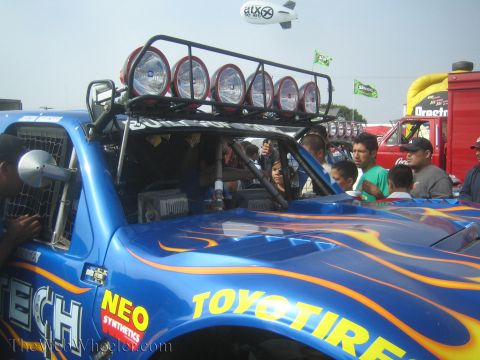 1990 Ford Bronco - blue off-road race truck with orange flame paint, a roof light bar, and a 'Toyo Tires' decal draws spectators under a cloudy sky.