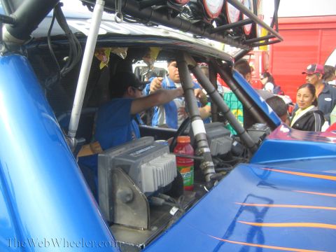 1990 Ford Bronco - open cockpit of a blue flame-painted off-road race truck reveals its roll cage, wiring, and dash electronics as people work on it.