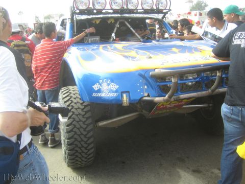 1990 Ford Bronco - front of a blue and yellow off-road race truck with a roof light bar and PCI Race Radios decal is surrounded by a crowd.