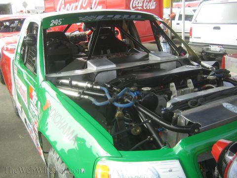 1990 Ford Bronco - green off-road race truck '245' sits with its hood removed, exposing the engine, plumbing, and roll cage, a Coca-Cola truck behind.