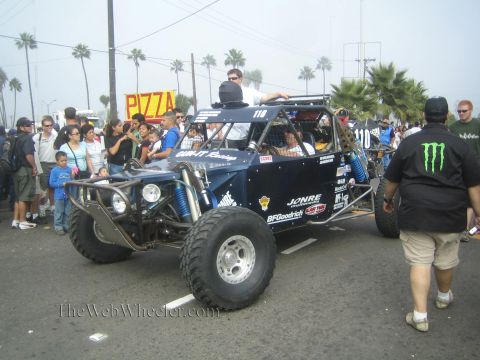 1990 Ford Bronco - dark blue open-frame off-road buggy marked '110' drives through a crowded palm-lined street past a Monster-shirted spectator.