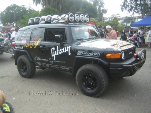 1990 Ford Bronco - black Toyota FJ Cruiser race rig with a roof light bar, stacked helmets, and Gibson and BFGoodrich decals parks among spectators.