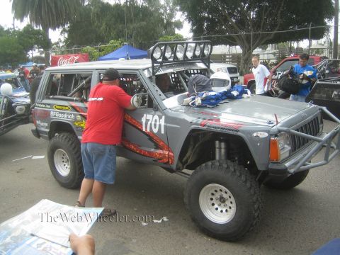 1990 Ford Bronco - crewman leans into the cab of a gray and red number 1701 off-road race Jeep Cherokee at a desert race staging area.
