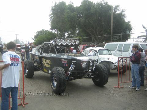 1990 Ford Bronco - black single-seat desert race buggy with Corona Extra livery and a four-light roof bar sits among spectators at a race staging area.