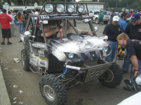 1990 Ford Bronco - black and silver Yamaha Rhino race side-by-side numbered 1818 with a roof light bar surrounded by crew at a race event.