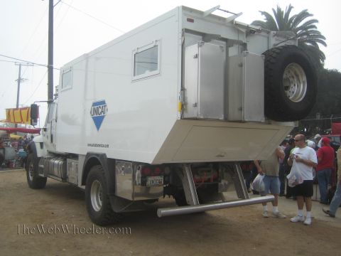 1990 Ford Bronco - Rear quarter view of a large white Unicat 4x4 expedition camper truck with a rear-mounted spare, spectators standing nearby.