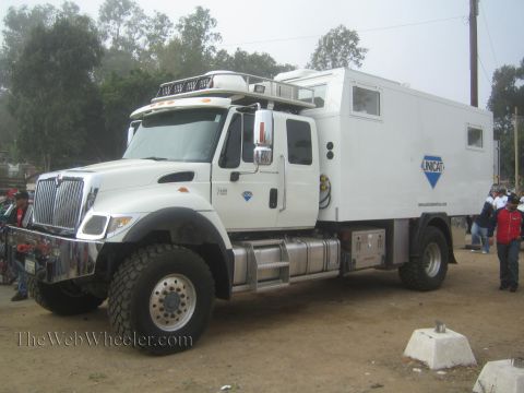 1990 Ford Bronco - large white International 4x4 chassis Unicat expedition camper truck parked on a dirt lot at a race gathering.