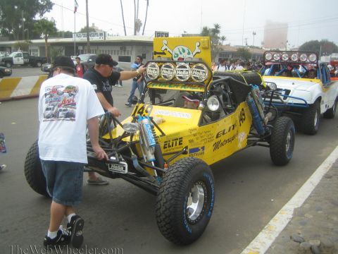 1990 Ford Bronco - yellow tube-frame desert race buggy numbered 149 with multiple Hella lights, crew gathered around at staging.