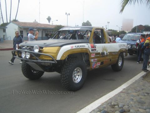 1990 Ford Bronco - gold and white single-cab prerunner race truck with sunburst graphics and dual round headlights moving down a street.
