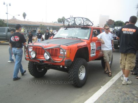 1990 Ford Bronco - orange lifted Jeep Cherokee race vehicle numbered 1702 with a roof light bar and tube bumper among crew at staging.