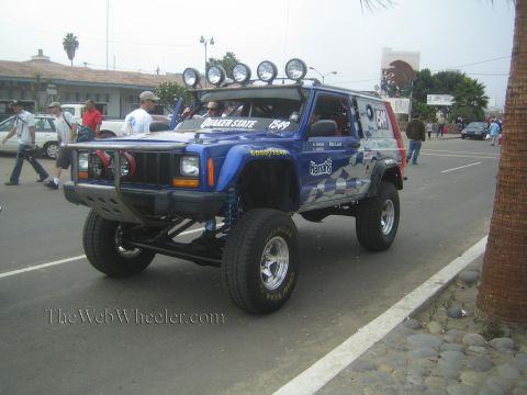 1990 Ford Bronco - blue lifted Jeep Cherokee race vehicle numbered 1549 with Quaker State graphics and a roof light bar on a street.