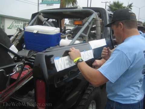 1990 Ford Bronco - man in a blue shirt applies a West Coast vinyl decal to the side of a black off-road race truck loaded with a cooler.