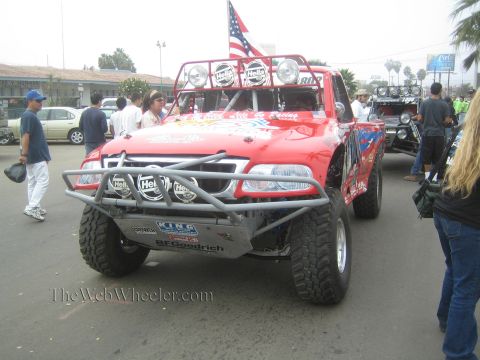 1990 Ford Bronco - red tube-bumper off-road race truck flying an American flag idles on a street as spectators look on.