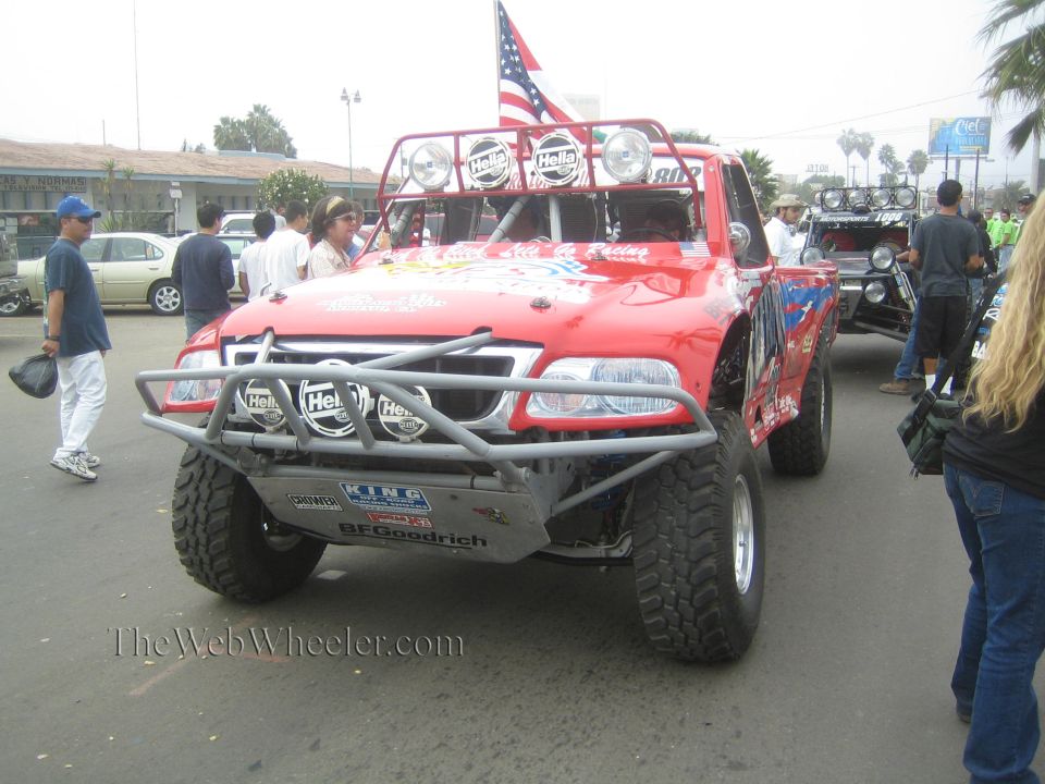 Red off-road race truck with light bar and US flag at a street gathering