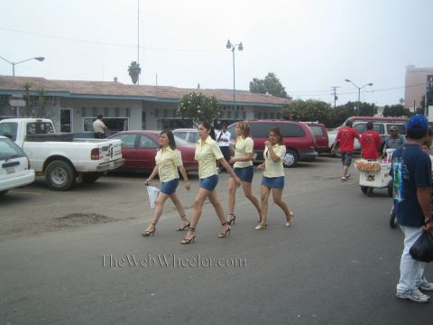 1990 Ford Bronco - Four women in matching yellow tops and short denim skirts walk across a dusty parking lot near parked trucks.