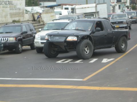 1990 Ford Bronco - black Ford extended-cab pickup with raised off-road suspension drives down a street painted with 'ALTO' lane markings.