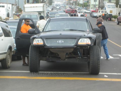 1990 Ford Bronco - black Ford pickup with exposed long-travel front suspension sits in traffic as bystanders look at it.