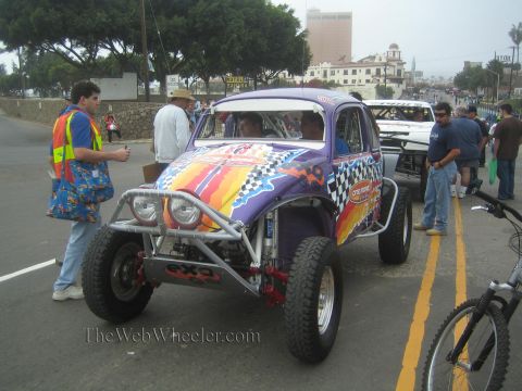 1990 Ford Bronco - purple Baja Bug with flame and checkered graphics sits roadside as a vest-clad official and onlookers gather.