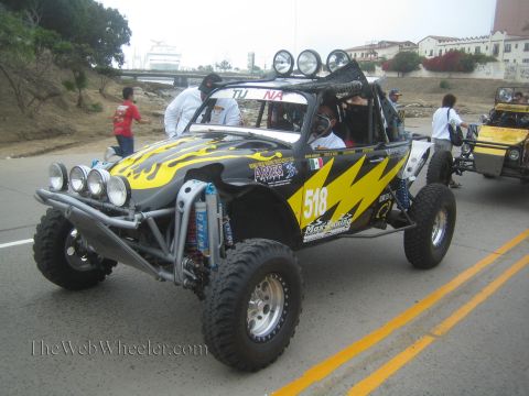1990 Ford Bronco - black and yellow number 518 tube-frame off-road buggy carrying a Mexican flag drives along a paved road.
