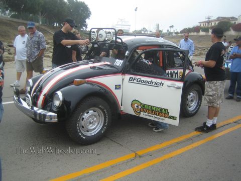 1990 Ford Bronco - black-and-white VW Beetle Baja race car number 1101 with BFGoodrich livery is surrounded by spectators on a road.