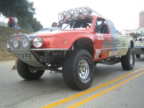 1990 Ford Bronco - red off-road race truck with a roof-mounted light bar and tube bumper sits parked on a sloped street.