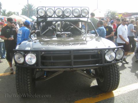 1990 Ford Bronco - raw unpainted off-road race truck with a six-light roof bar and tube bumper sits in a crowd at an event.