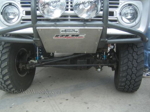 1990 Ford Bronco - Close-up of an off-road truck's front end showing Lightforce lamps, a skid plate and exposed steering linkage.