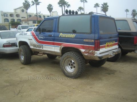 1990 Ford Bronco - lifted blue Ford SUV with Goodyear and Pit Crew livery and roof lights sits on dirt among parked vehicles near palm trees.