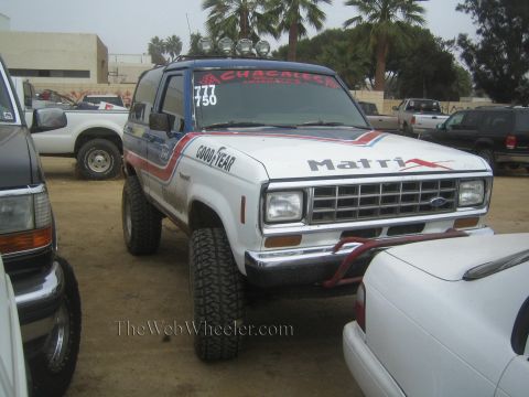 1990 Ford Bronco - white lifted Ford SUV marked Matrix and Goodyear with race numbers and roof-mounted lights parked on dirt.