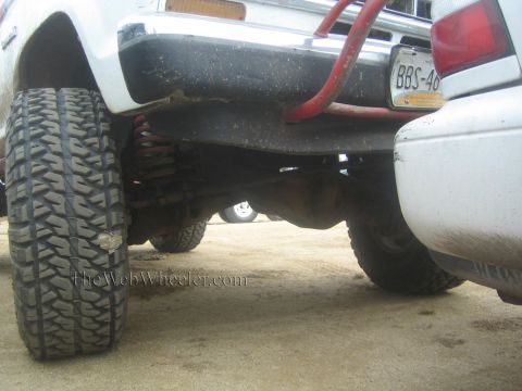 1990 Ford Bronco - low under-vehicle shot of a lifted white truck reveals the coil-spring suspension, oversized mud tire and a license plate.
