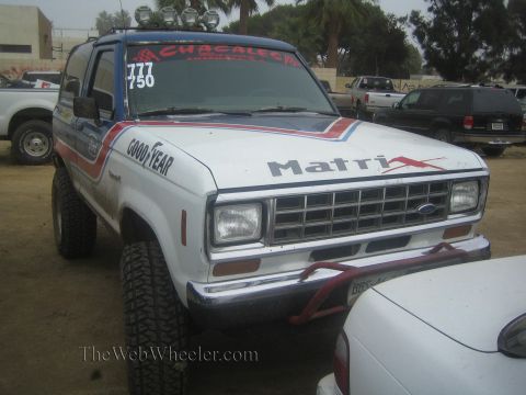 1990 Ford Bronco - white lifted Ford SUV with Matrix livery, roof lights and race numbers sits among parked vehicles on a dirt lot.