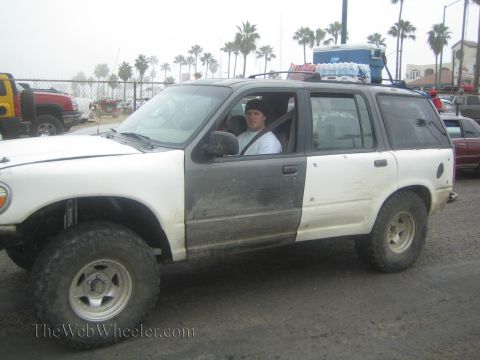 1990 Ford Bronco - muddy white Ford Explorer with lift kit and roof rack holding water bottles, a young driver visible through the window.