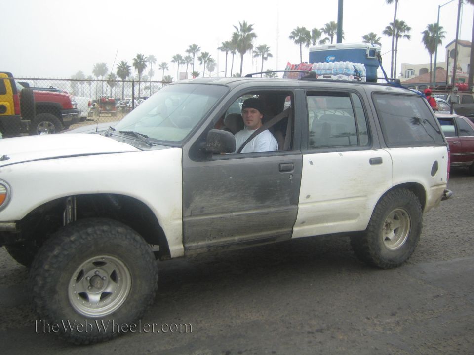 White Ford Explorer with lift and mud tires, driver seated inside, roof rack with water jugs