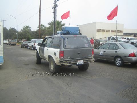 1990 Ford Bronco - white lifted SUV with roof-mounted gear drives down a street lined with red flags and parked cars under an overcast sky.