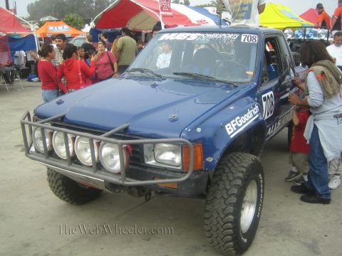 1990 Ford Bronco - blue off-road race truck numbered 708 with a tubular bumper and four round driving lights surrounded by spectators.