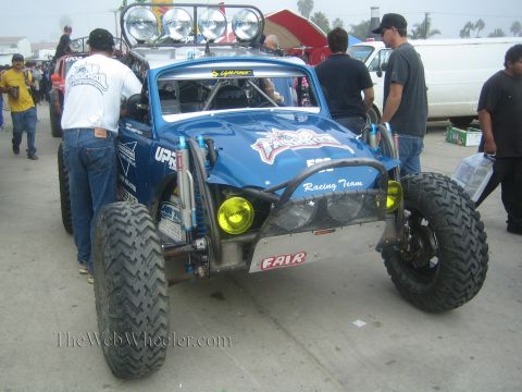 1990 Ford Bronco - blue tube-chassis off-road race vehicle with long-travel suspension and yellow lights surrounded by crew in a pit area.