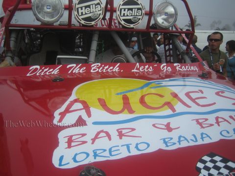 1990 Ford Bronco - Close-up of a red race vehicle's hood lettered Augies Bar and Lets Go Racing, topped by Hella driving lights on a roll cage.