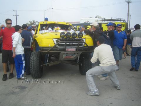 1990 Ford Bronco - yellow Ford off-road race truck numbered 719 with HID lights and tube bumper is pushed through a crowd at staging.