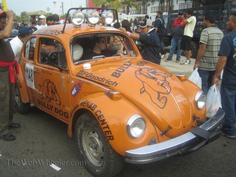 1990 Ford Bronco - orange Volkswagen Beetle race car numbered 1148 with Bully Dog and roof lights is surrounded by spectators on a street.