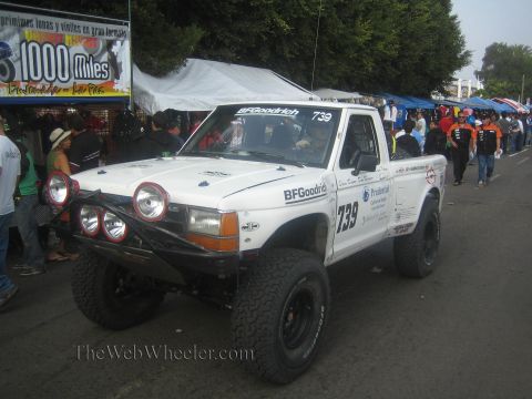 1990 Ford Bronco - white Ford prerunner race truck numbered 739 with round driving lights and BFGoodrich decals parked at a busy street event.