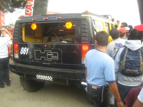 1990 Ford Bronco - rear of a black Hummer H2 numbered 861 with amber lights and Checkers decals is crowded by onlookers.