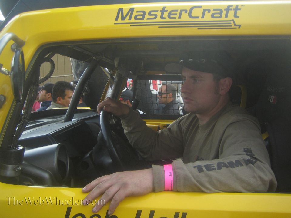Man in green shirt and cap seated at wheel inside yellow race vehicle with MasterCraft cage
