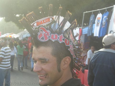 1990 Ford Bronco - smiling man sports a tall novelty mohawk hat covered in Maxxis and Fotos logos amid an outdoor vendor crowd.