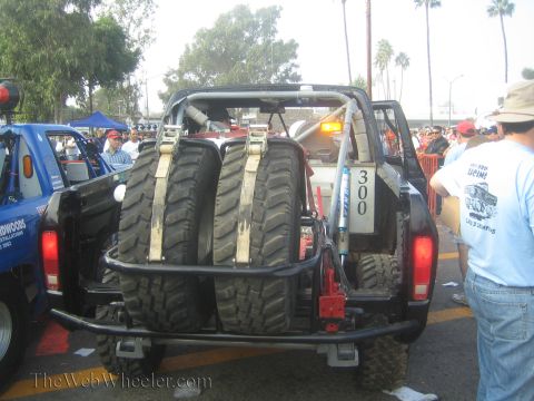 1990 Ford Bronco - rear of a tube-frame off-road race truck numbered 300 carries two strapped spare mud tires among spectators.