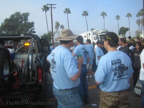 1990 Ford Bronco - Spectators in matching light blue Moss Bros Racing T-shirts gather beside a race truck under palm trees.