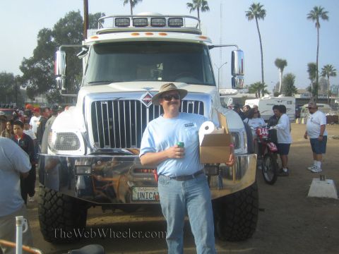 1990 Ford Bronco - man in a blue shirt and hat holds cans in front of a large chrome International chase truck with roof lights on a dirt lot.