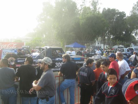1990 Ford Bronco - crowd of spectators stands behind a barrier as off-road race trucks including a Bronco are staged in a hazy dirt lot.